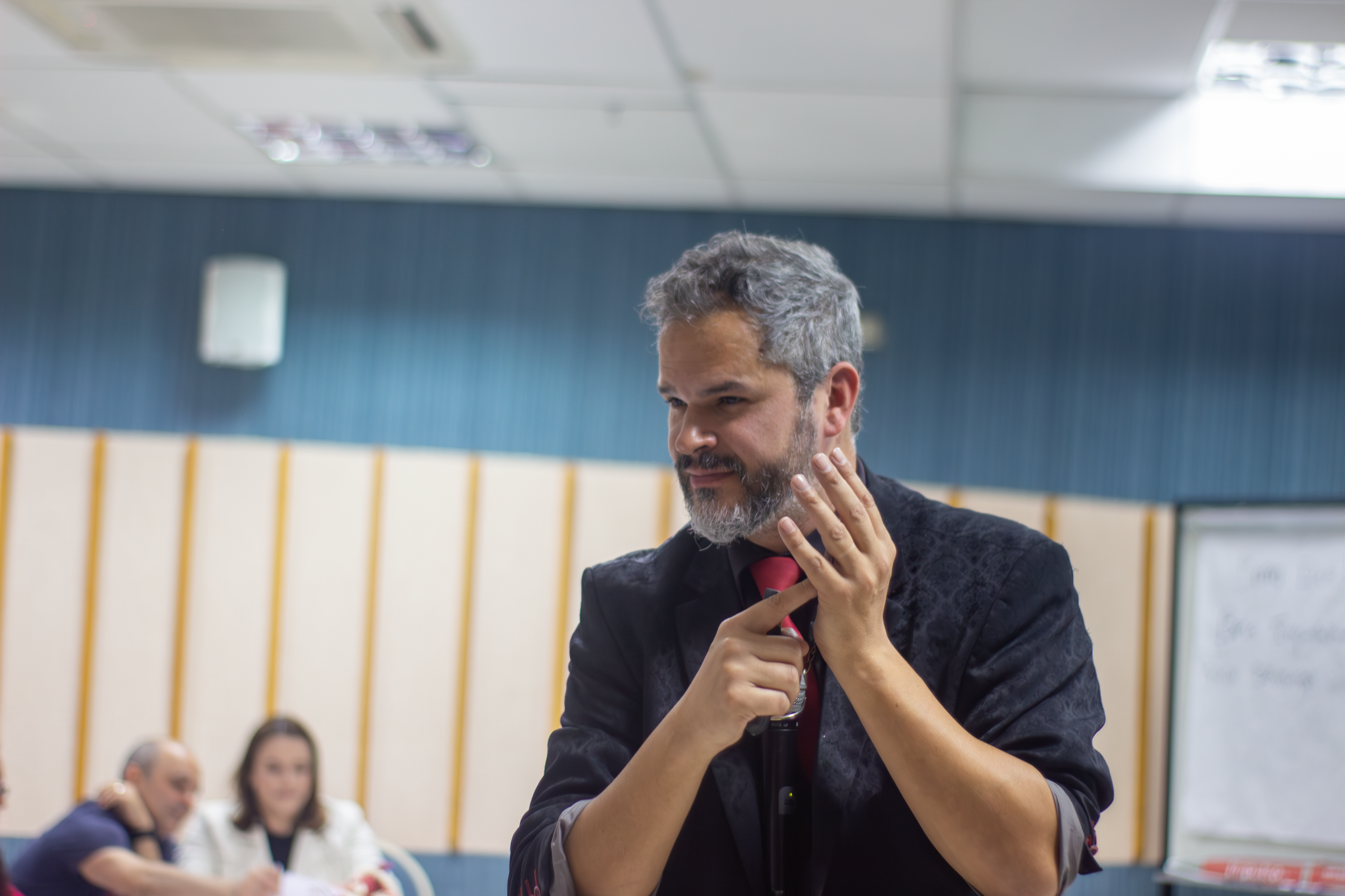 Homem de barba grisalha, usando paletó preto e gravata vermelha, em gesto expressivo durante uma apresentação em ambiente corporativo, com pessoas ao fundo observando.