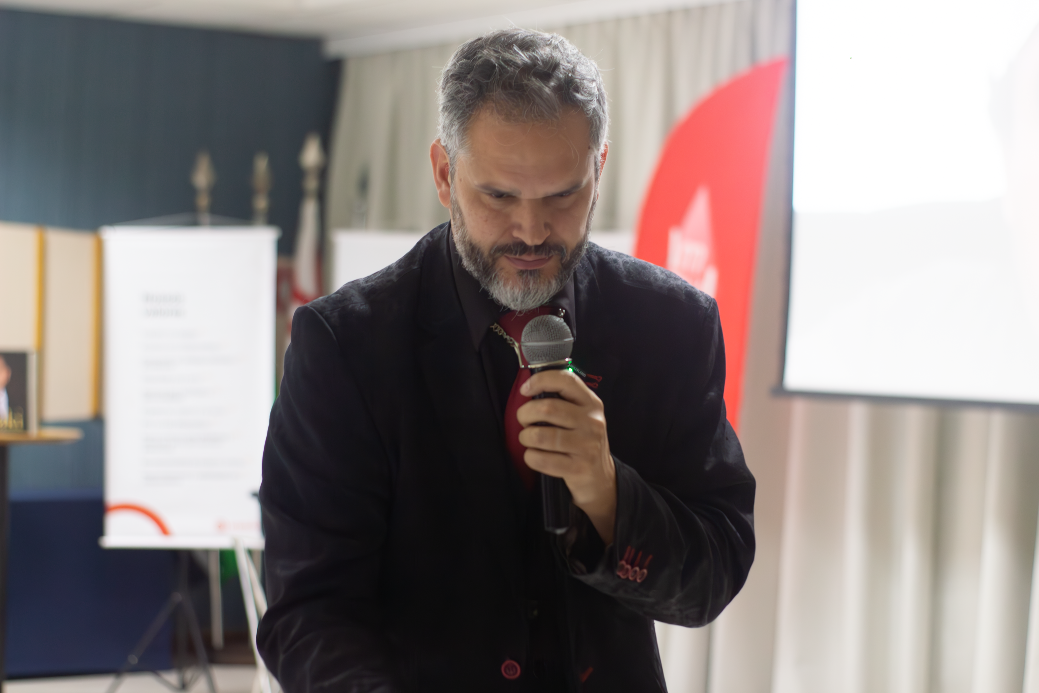 Homem de barba grisalha e terno preto com camisa vermelha segura um microfone durante uma apresentação em auditório corporativo.