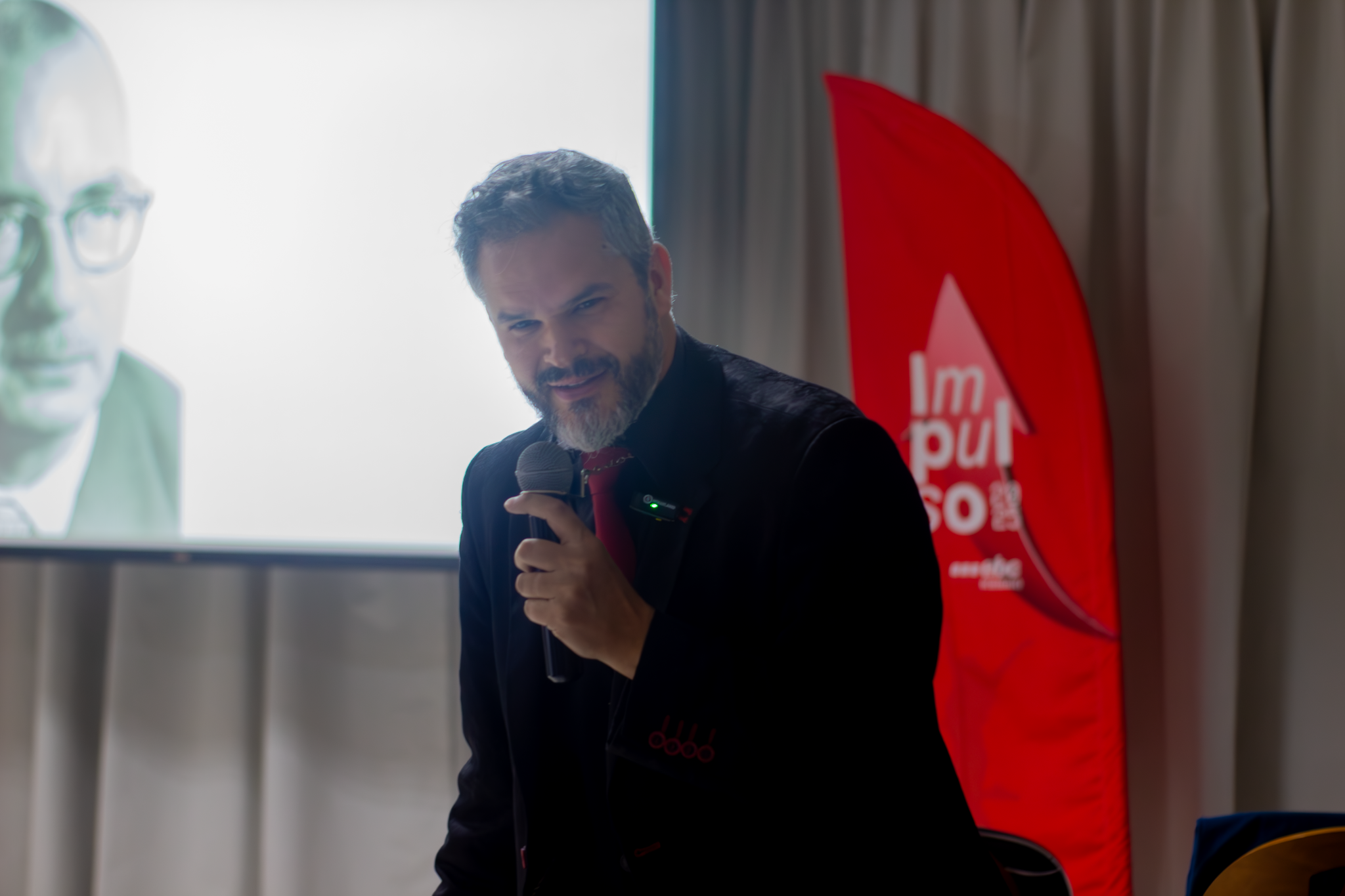 Palestrante segurando microfone durante apresentação corporativa, com projeção em tela e banner vermelho ao fundo.