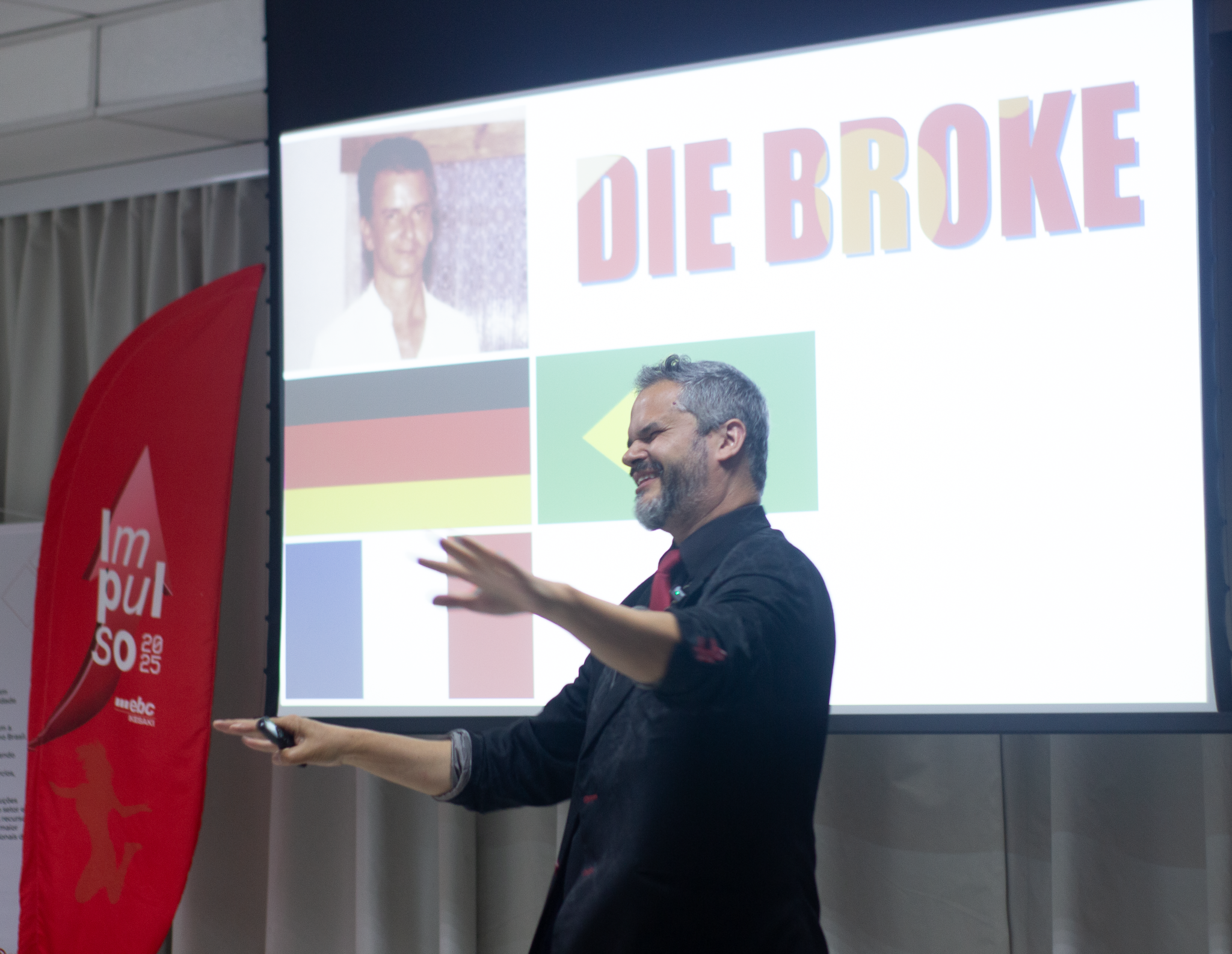 Palestrante motivacional de meia-idade em momento energético no palco, falando para uma plateia corporativa durante um evento de fim de ano.