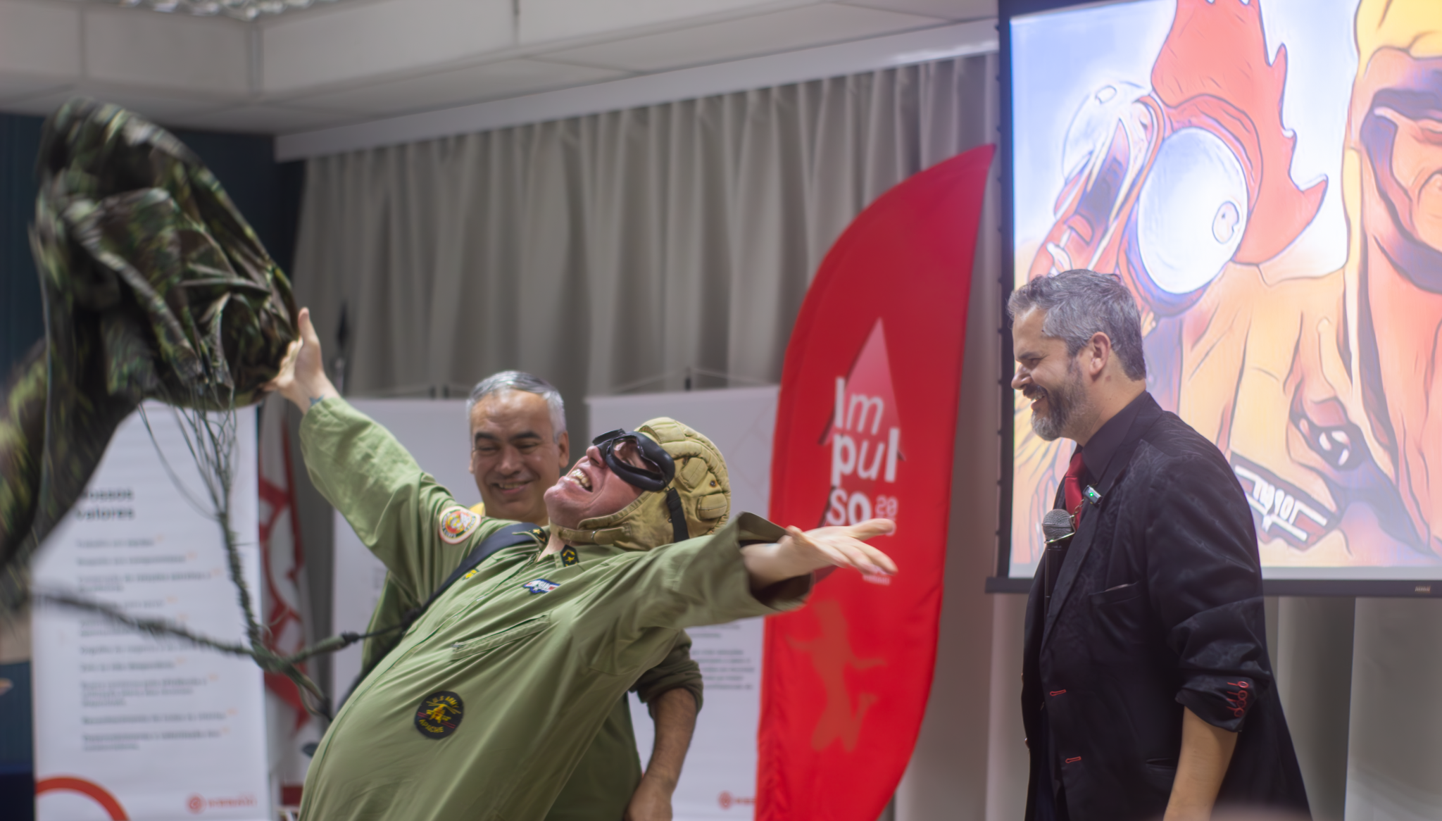 Homem fantasiado de aviador abre os braços em expressão de alegria durante uma palestra interativa, enquanto o palestrante e outro participante sorriem ao lado, em um ambiente corporativo animado.