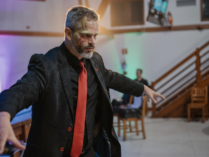Homem caracterizado como zumbi corporativo, usando terno escuro com gravata vermelha, fazendo pose cômica em um ambiente de palestra.