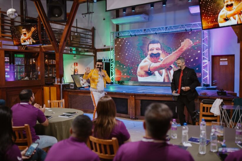 Palestrante Paul & Jack interage com o público durante palestra motivacional, com artista caracterizado como Freddie Mercury em apresentação temática de fim de ano.