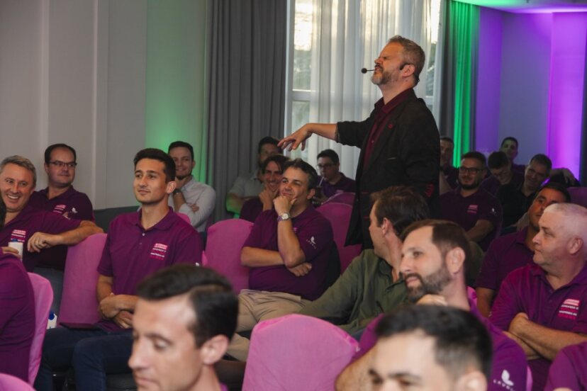 Palestrante com microfone de cabeça interage sorrindo com a plateia em um treinamento corporativo; participantes usam camisetas roxas, cadeiras com capas magenta, iluminação ambiente em tons de verde e roxo, clima descontraído.