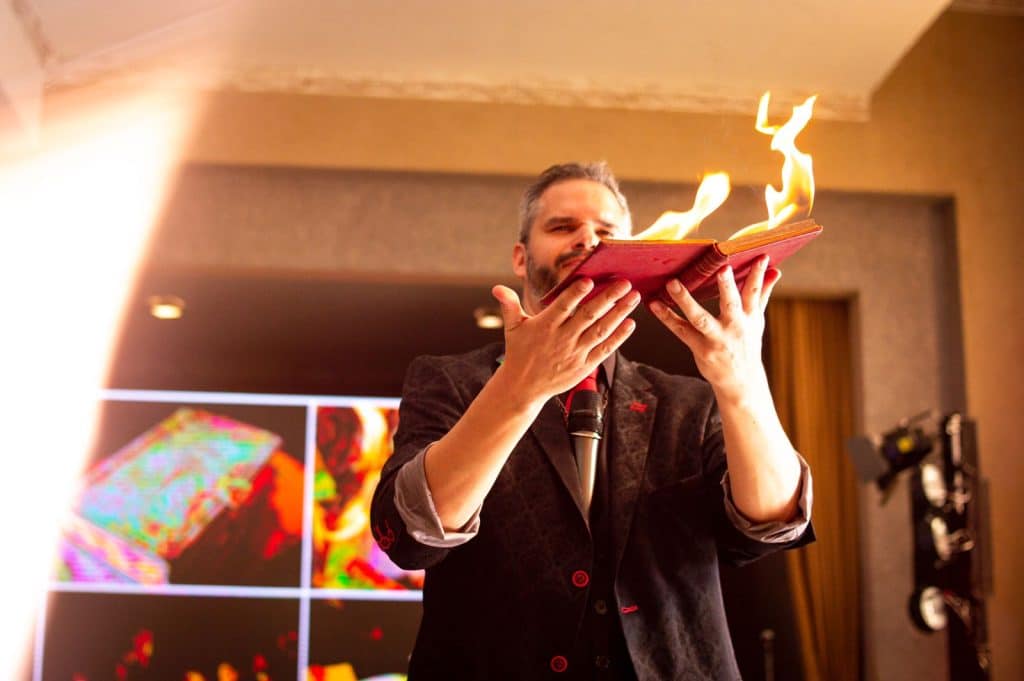 Mágico se apresenta segurando um livro em chamas durante uma palestra-show, vestindo terno escuro com detalhes vermelhos, em um ambiente corporativo iluminado.