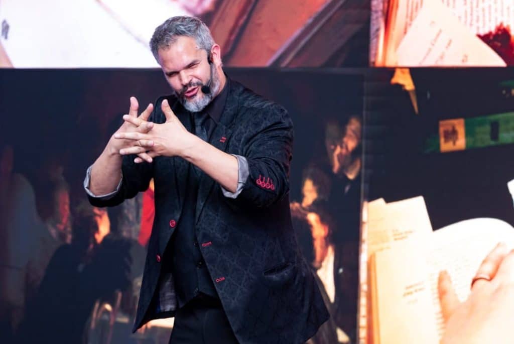 Mágico Paul & Jack em apresentação solo, realizando truque com as mãos no palco, usando terno preto com detalhes vermelhos e microfone headset.