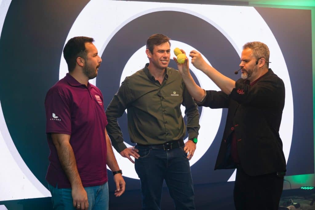 Mágico Paul & Jack realiza truque com esponjas amarelas no palco, interagindo com dois participantes durante evento corporativo.
