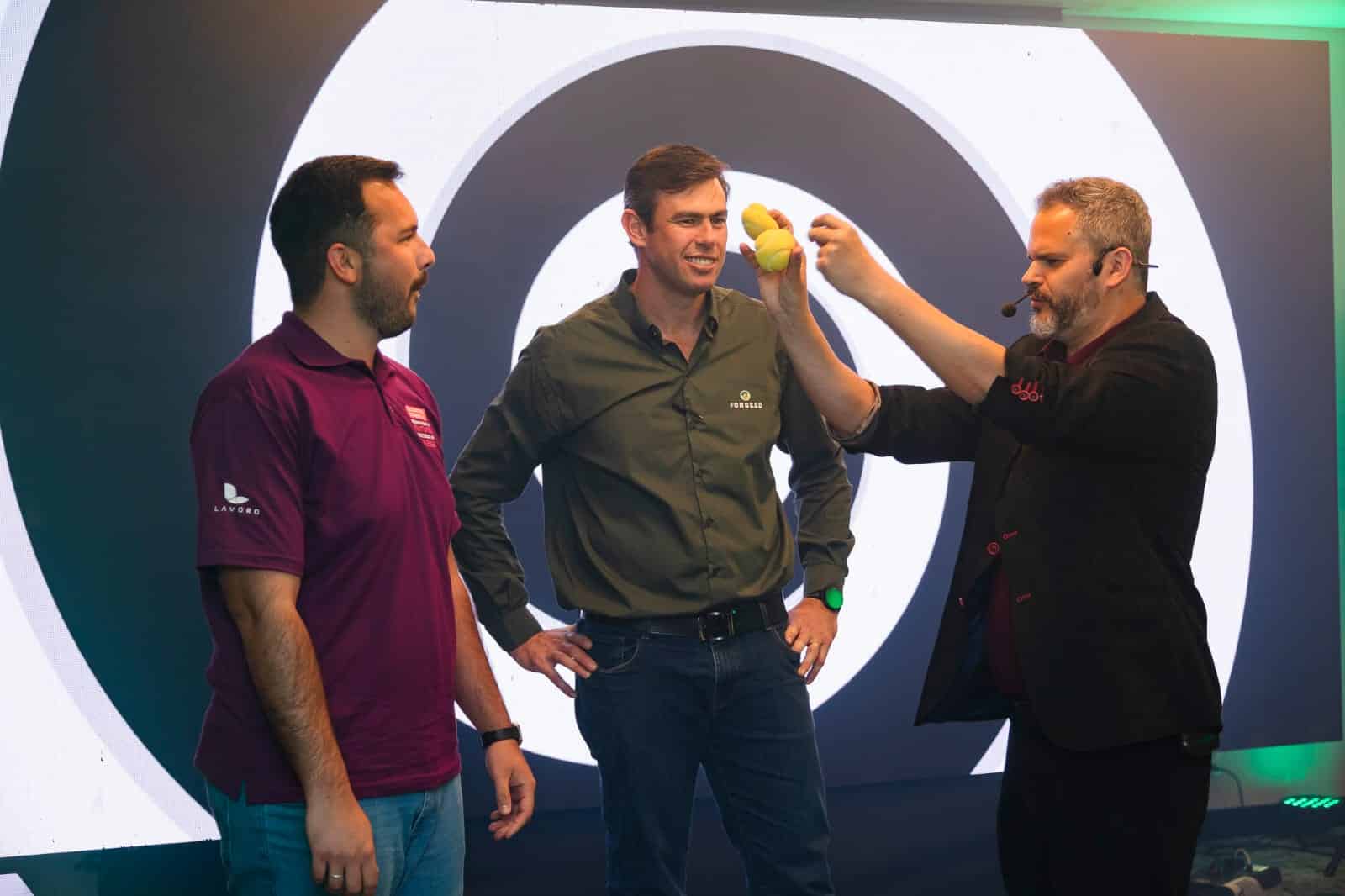 Mágico Paul & Jack realiza truque com esponjas amarelas no palco, interagindo com dois participantes durante evento corporativo.