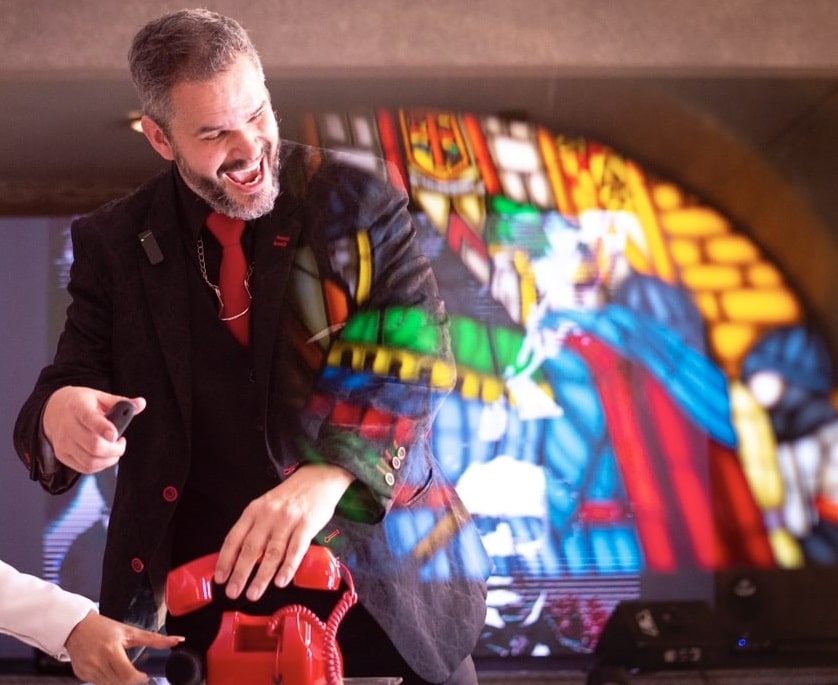 Mágico Paul & Jack sorri enquanto realiza truque com telefone vermelho, com vitral colorido ao fundo refletindo luz sobre seu paletó.