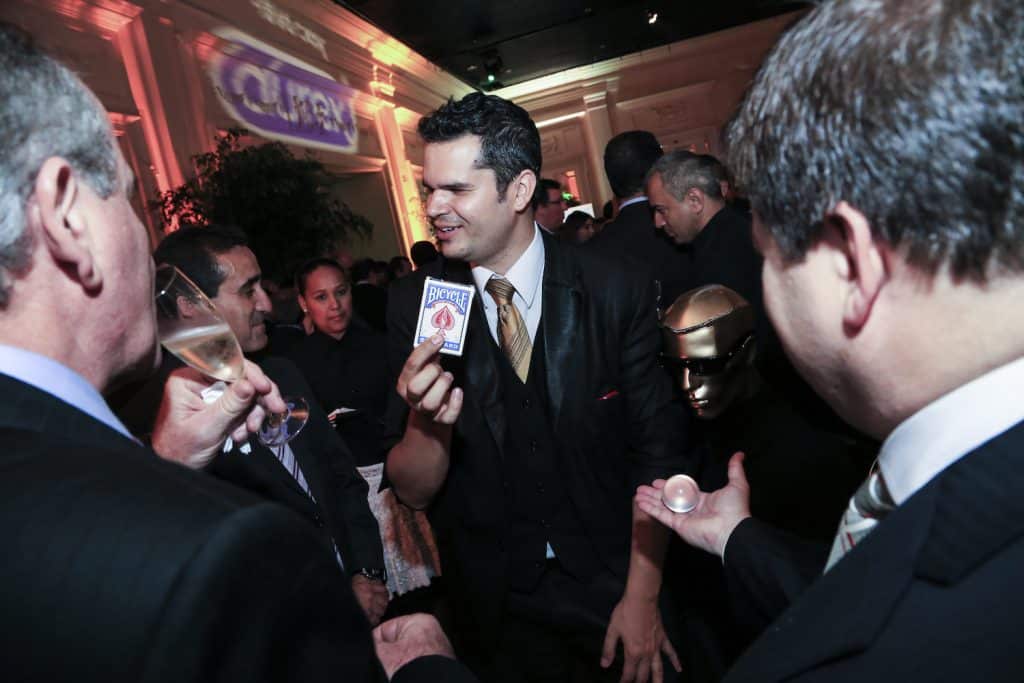 Mágico Paul & Jack realizando truque com baralho em evento corporativo, cercado por convidados em trajes sociais atentos à apresentação.