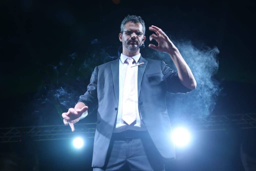Homem em cena, usando terno escuro e microfone de cabeça, iluminado por luzes fortes e envolto por fumaça cênica, realizando um gesto expressivo com as mãos durante uma apresentação de mágica no palco.