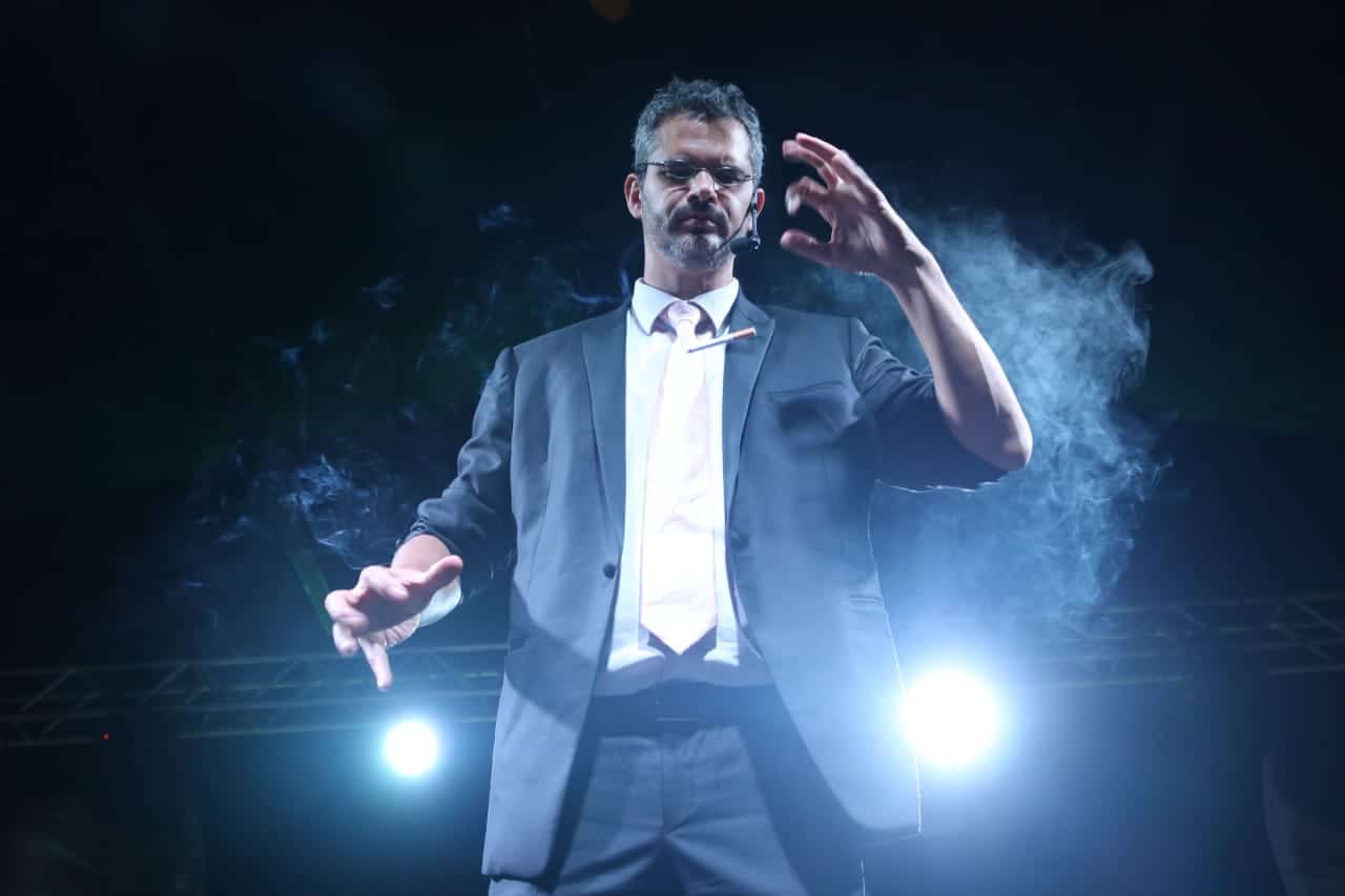 Homem em cena, usando terno escuro e microfone de cabeça, iluminado por luzes fortes e envolto por fumaça cênica, realizando um gesto expressivo com as mãos durante uma apresentação de mágica no palco.