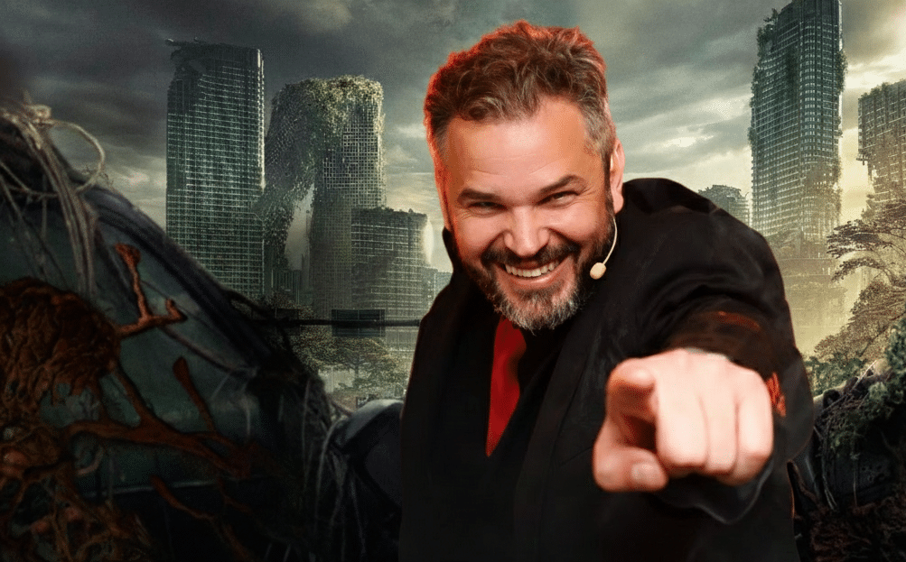 Homem sorridente, de frente para a câmera, apontando diretamente para o espectador, usando microfone de lapela, com cenário urbano pós-apocalíptico ao fundo, prédios destruídos e cobertos por vegetação, em referência visual ao universo de The Last of Us.