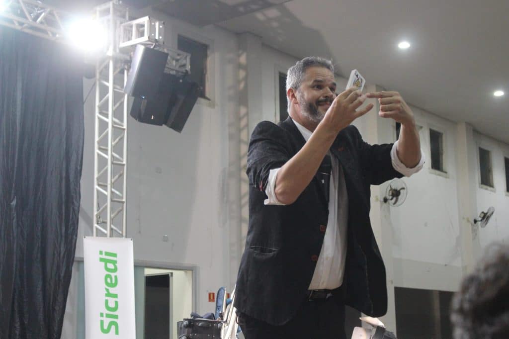 Palestrante em palco segurando uma carta de baralho durante apresentação, usando gesto visual para ilustrar um conceito diante do público.