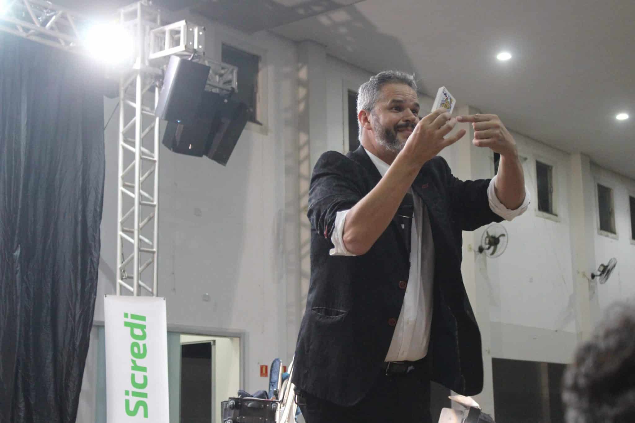 Palestrante em palco segurando uma carta de baralho durante apresentação, usando gesto visual para ilustrar um conceito diante do público.