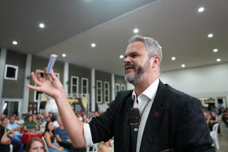 Homem palestrando em auditório lotado, segurando um objeto pequeno na mão enquanto fala ao público, com microfone e expressão engajada.