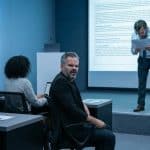 Apresentador lê um documento diante de público desatento em sala moderna; alguns bocejam, usam celular ou demonstram tédio, enquanto tela exibe slide denso e pouco legível.