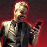 Homem sorridente vestindo terno escuro e gravata, olhando para a tela do celular com expressão de surpresa positiva, sobre fundo vermelho com efeito de destaque ao redor do corpo.