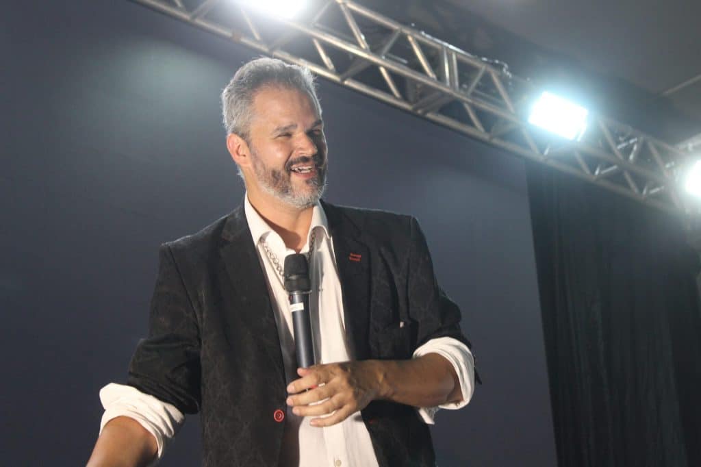 Palestrante segurando microfone no palco, sorrindo, durante apresentação para público em ambiente corporativo com iluminação profissional.