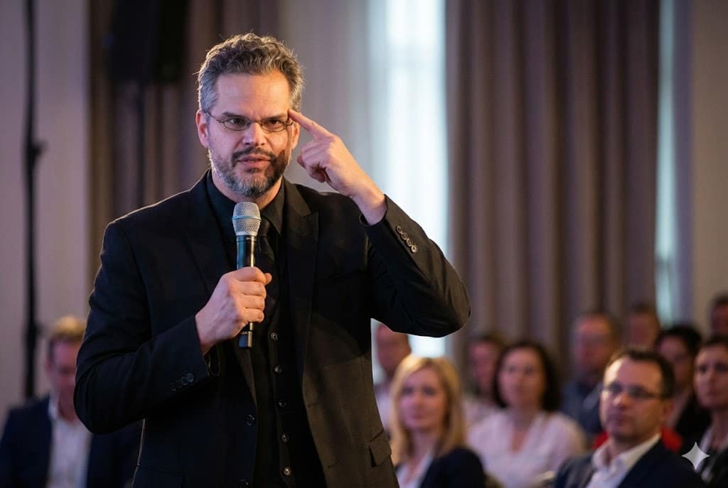 Homem de barba e óculos em um palco, segurando um microfone e apontando o dedo indicador para a própria têmpora enquanto fala para uma plateia.