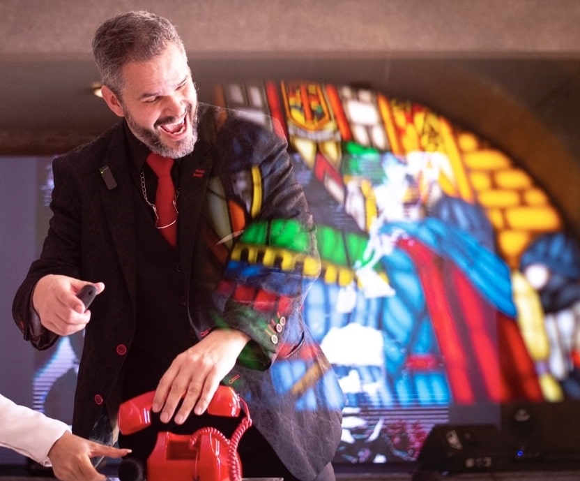Homem sorridente em terno preto com detalhes vermelhos, gesticulando ao lado de um telefone vermelho diante de um vitral colorido com figura medieval.