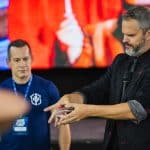Homem de jaqueta preta com detalhes vermelhos e microfone de lapela realiza truque de cartas diante de plateia; outro homem de camiseta azul observa atentamente, com tela colorida ao fundo.