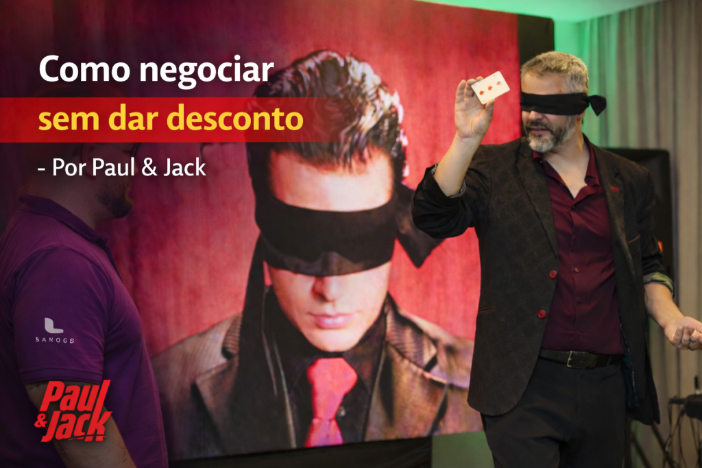 Capa de blog 16:9 com Paul à direita, vendado, segurando uma carta de baralho e usando terno escuro com camisa vinho. Fundo desfocado com telão vermelho e imagem de um homem vendado. À esquerda, texto grande: “Como negociar sem dar desconto: confiança, tempo e tática para fechar acordos melhores – Com Paul & Jack”, com faixa vermelha destacando “sem dar desconto”. Logo “Paul & Jack” no canto inferior esquerdo.