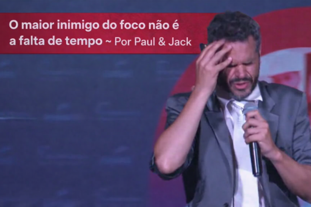Paul aparece no palco, segurando um microfone e levando a mão à testa, em uma expressão de tensão e concentração. Na parte superior da imagem, uma faixa vermelha destaca o título: “O maior inimigo do foco não é a falta de tempo ~ Por Paul & Jack”.