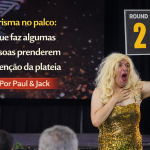 Apresentador no palco com vestido dourado segurando placa “Round 2”, com o título “Carisma no palco: o que faz algumas pessoas prenderem a atenção da plateia – Por Paul & Jack”.