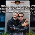 Montagem em formato 16:9 inspirada em pôster cinematográfico, mostrando Paul & Jack abraçando Arnold Schwarzenegger caracterizado como o Exterminador do Futuro. Os dois aparecem ao centro, em frente a um painel metálico vertical, com fundo de palco escuro, iluminação azul e vermelha nas laterais e telas desfocadas ao fundo. Na parte superior da imagem, há um título grande em estilo metálico: “Os 3 segredos de sucesso de Arnold Schwarzenegger que mudaram minha vida”, seguido de “- por Paul & Jack”. No topo, aparece um selo circular que remete à estética de capa de DVD.